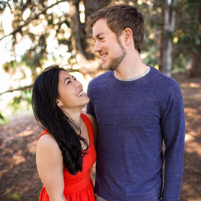 Melody laughing while wearing orange dress and facing Alex smiling wearing a purple shirt. Forest of evergreen trees in the background